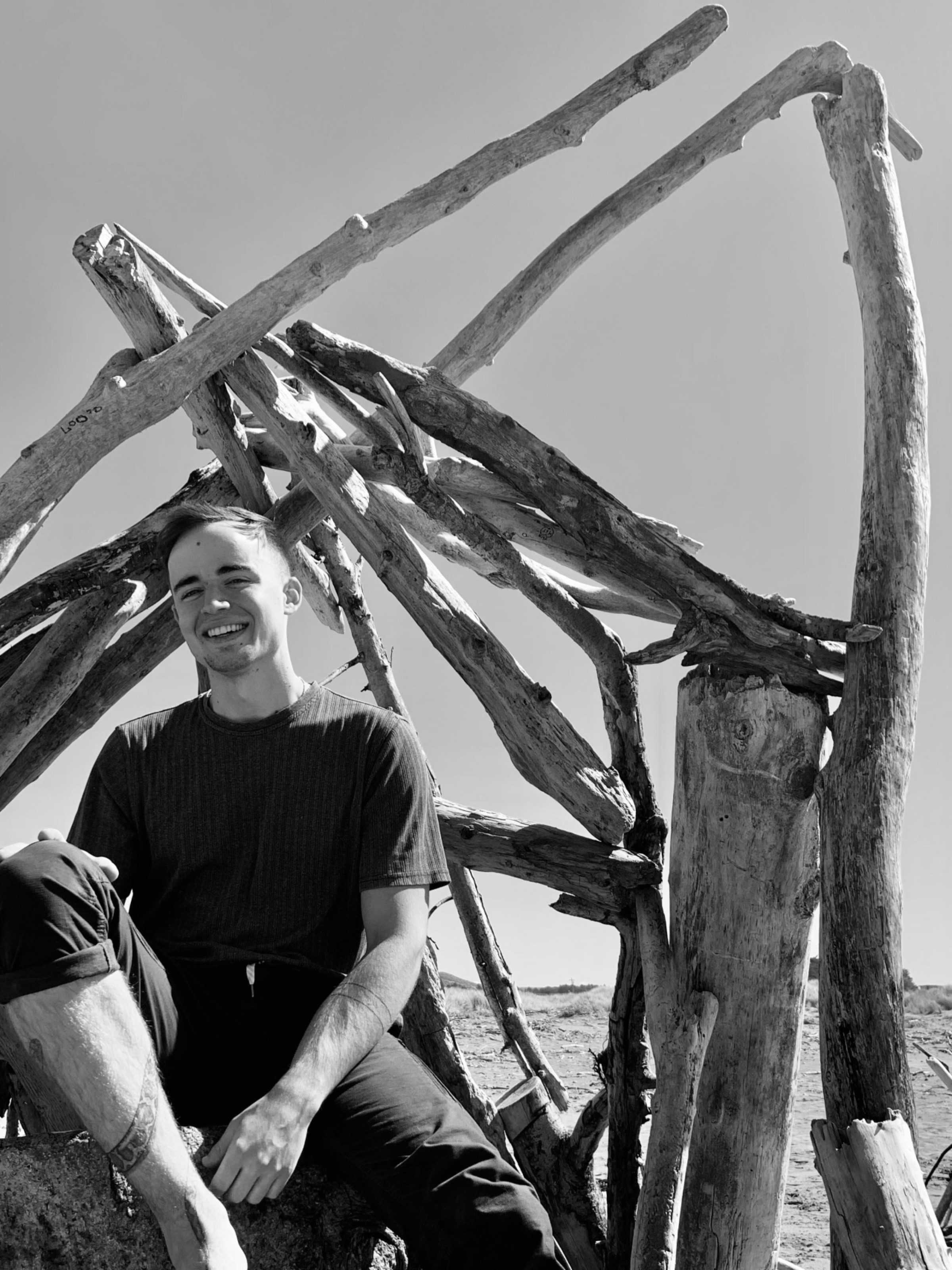Thor Nolan smiling, sitting in a house made of driftwood on a beach